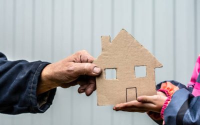 A man's hand holds a paper house cut out of cardboard. Nearby, children's hands hold the house from below.